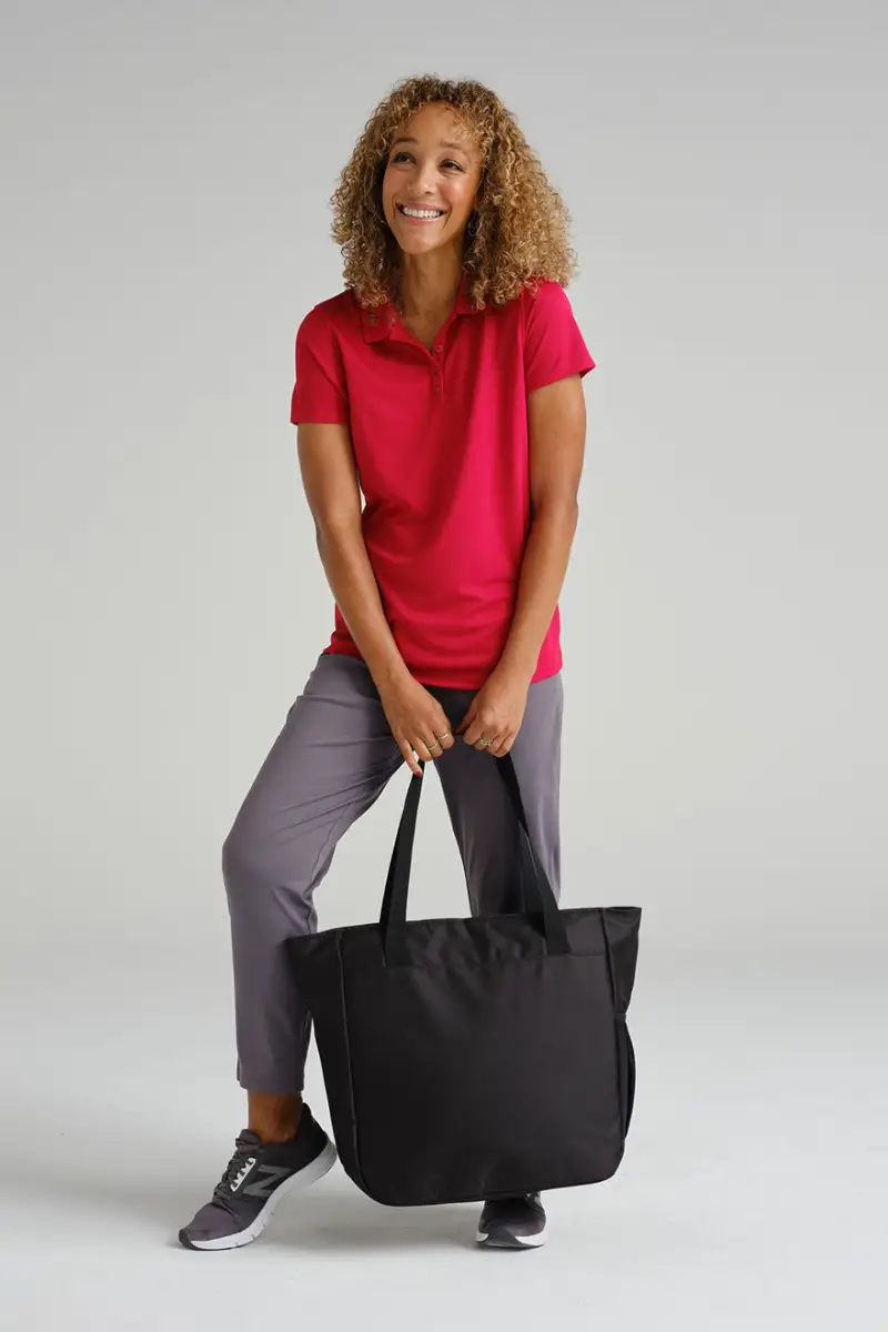 A woman stands with a black tote bag, wearing a red polo shirt and gray pants, with a casual athletic style.