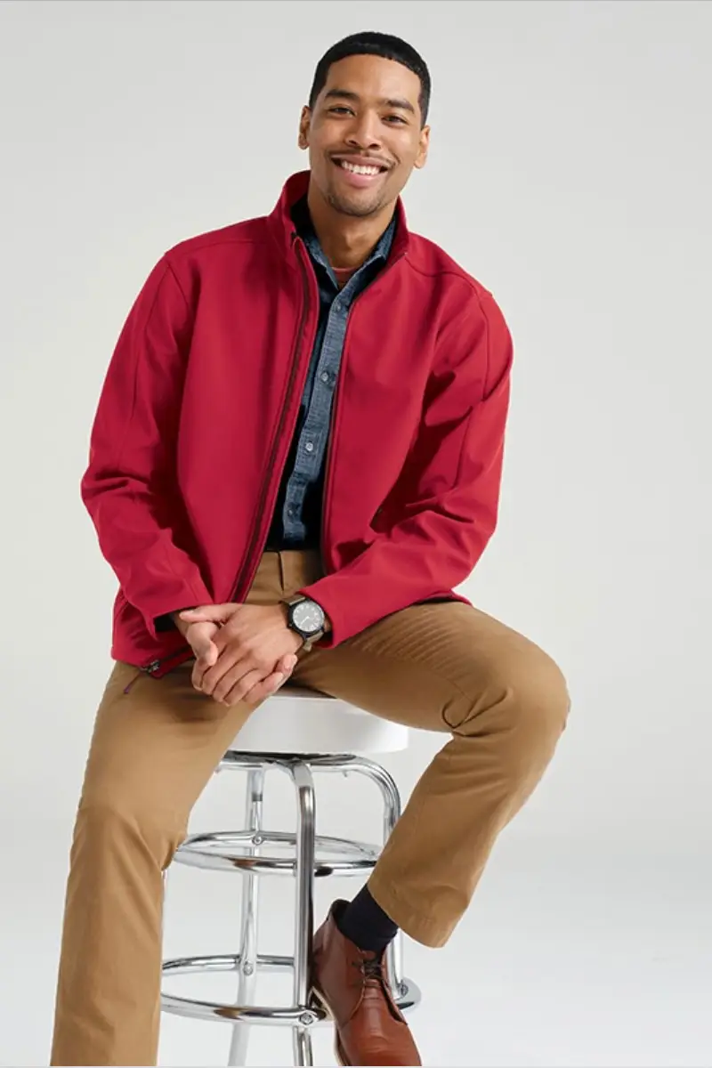 A man sits on a chrome stool, wearing a red jacket, denim shirt, tan pants, and brown shoes, exuding a casual, stylish vibe.