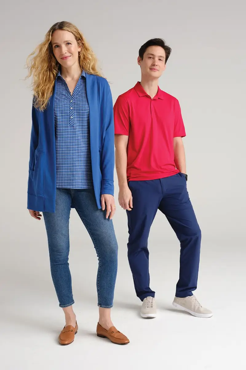 A woman in a blue cardigan over a checkered top and jeans stands beside a man in a red polo and navy pants, both stylishly dressed.