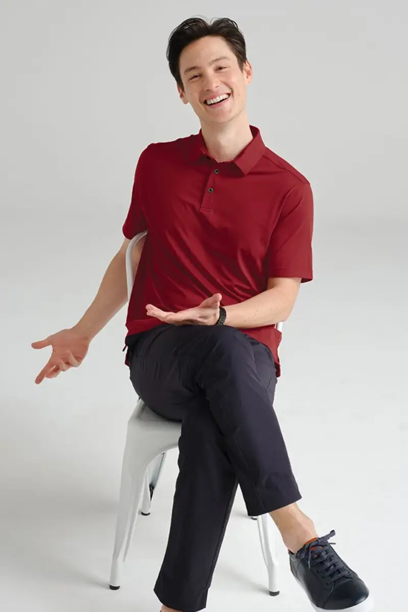 A person in a red polo shirt sits casually on a chair, gesturing with their hands while wearing dark pants and sneakers.